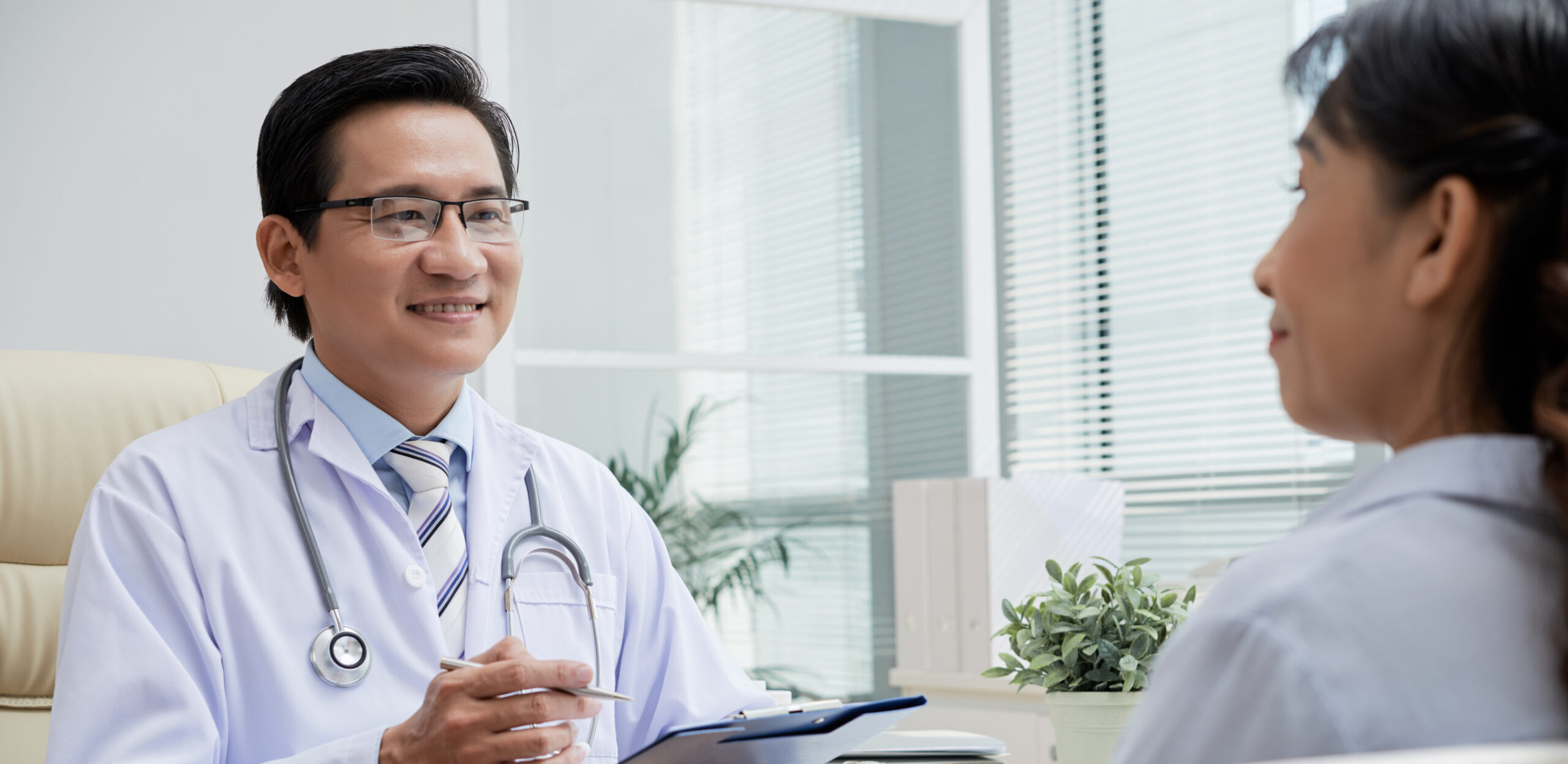 Friendly middle-aged physician wearing white coat sitting opposite senior patient and giving recommendations while having appointment at modern office